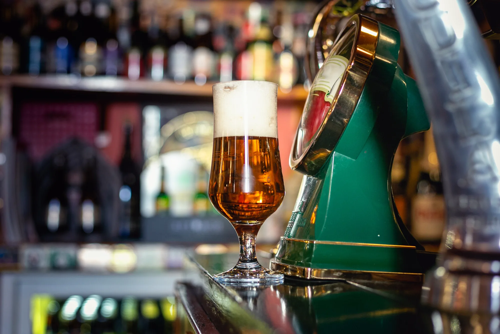 Copa de cerveza dorada con espuma sobre la barra, con las estanterías de botellas al fondo