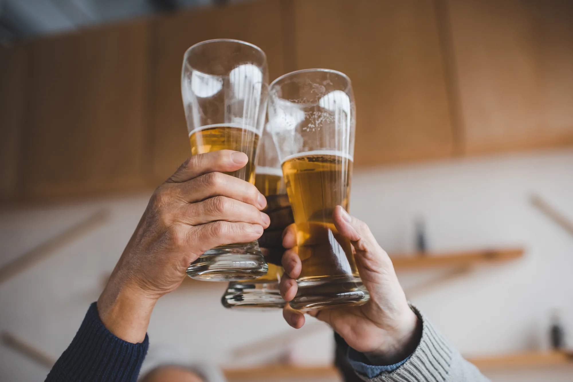 Dos manos brindan con vasos altos de cerveza sobre una mesa de madera clara