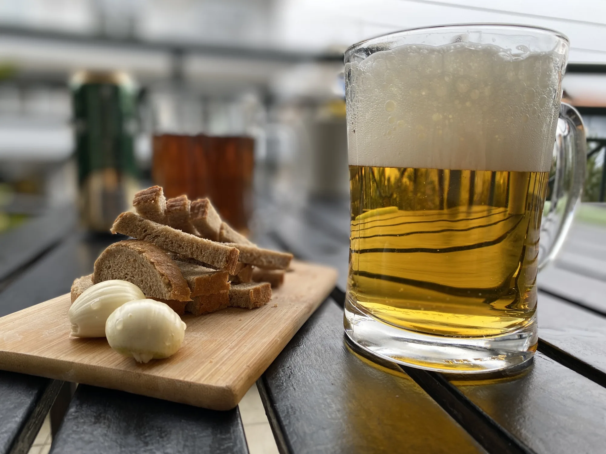 Un vaso alto de cerveza junto a una tabla de madera con pan y ajos, en una mesa de exterior
